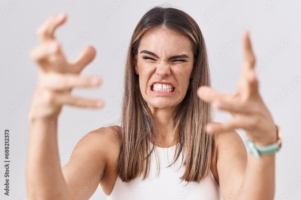 Sticker Hispanic young woman standing over white background shouting frustrated with rage, hands trying to strangle, yelling mad