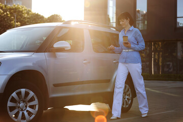 Elegant businesswoman in blue shirt, white pants holds paper cup of hot drink and smartphone while...