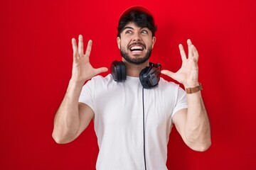 Hispanic man with beard wearing gamer hat and headphones crazy and mad shouting and yelling with...