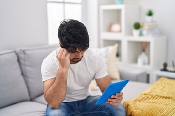 Hispanic man with beard using touchpad sitting on the sofa worried and stressed about a problem...