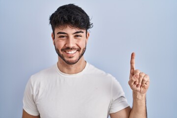 Hispanic man with beard standing over white background showing and pointing up with finger number one while smiling confident and happy. - obrazy, fototapety, plakaty