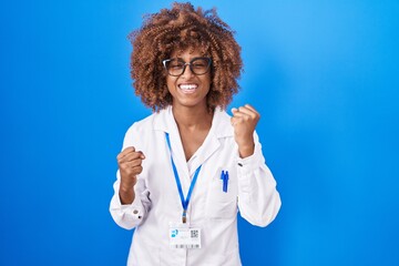 Young hispanic woman with curly hair wearing white coat and id card very happy and excited doing winner gesture with arms raised, smiling and screaming for success. celebration concept. - obrazy, fototapety, plakaty