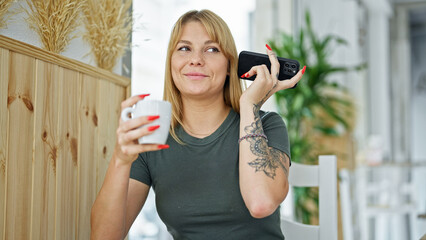 Young blonde woman listening to voice message by smartphone drinking coffee at coffee shop