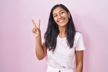 Young hispanic woman standing over pink background smiling with happy face winking at the camera...