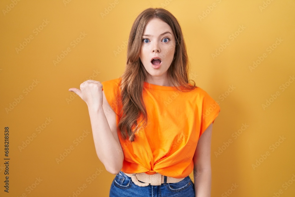 Poster caucasian woman standing over yellow background surprised pointing with hand finger to the side, ope