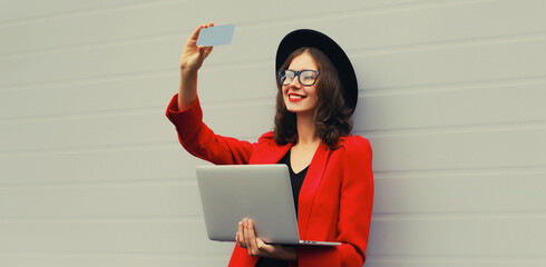 Businesswoman taking selfie with mobile phone working with laptop wearing red blazer jacket, eyeglasses, round hat on gray background, happy elegant woman holds pc computer