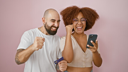 Beautiful couple standing together using smartphone and credit card celebrating over isolated pink...