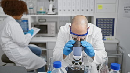 Naklejka premium Inside the buzzing lab, two committed scientists, man and woman, analysis partners, seated together, gloved hands busy writing on clipboard, eyes glued to the microscope in serious research