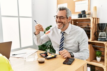 Middle age grey-haired man architect smiling confident eating sushi at office