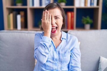 Middle age hispanic woman sitting on the sofa at home covering one eye with hand, confident smile on face and surprise emotion.