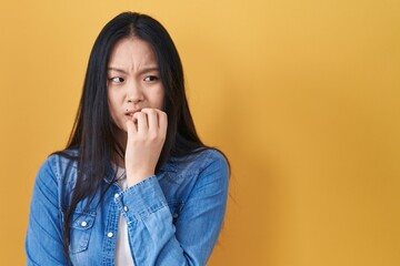 Young asian woman standing over yellow background looking stressed and nervous with hands on mouth biting nails. anxiety problem.