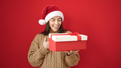 Young beautiful hispanic woman wearing christmas hat unpacking gift over isolated red background