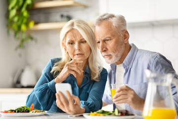 Concerned Senior Spouses Reading News On Smartphone During Breakfast In Kitchen