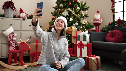 Obraz na płótnie Canvas Young beautiful hispanic woman make selfie by smartphone sitting on floor by christmas tree at home