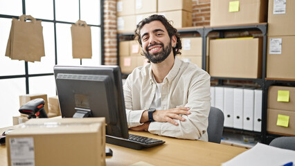 Young hispanic man ecommerce business worker using computer sitting on table with arms crossed...