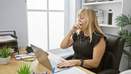 Beautiful middle age blonde woman, a professional business worker, intensely working on laptop at the office desk and coughing - an alarming sign of sickness - obrazy, fototapety, plakaty
