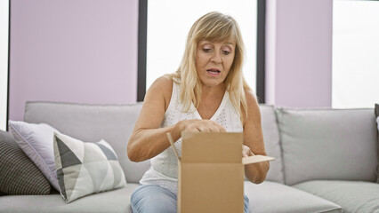 Beautiful middle age blonde woman, surprised while unpacking a cardboard box at home indoors,...
