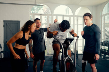 Multiracial young adults in sports clothing cheering on friend cycling on exercise bike at the gym - 688222758