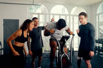 Tableaux sur verre Fitness Multiracial young adults in sports clothing cheering on friend cycling on exercise bike at the gym