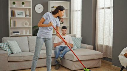 Beautiful couple expressing love, cleaning floor together at home. boyfriend casually texting on...
