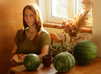 Fototapeta premium Selfie portrait of fair-haired woman in ethnic natural clothes in a country house, in the rays of the sun. What is sitting at the table on which there are large green watermelons.