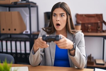 Hispanic woman working at the office playing video games in shock face, looking skeptical and...