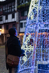 Latino man admires festive Christmas glow in Hondarribia's winter village square at dusk.
