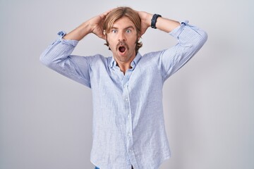 Caucasian man with mustache standing over white background crazy and scared with hands on head,...