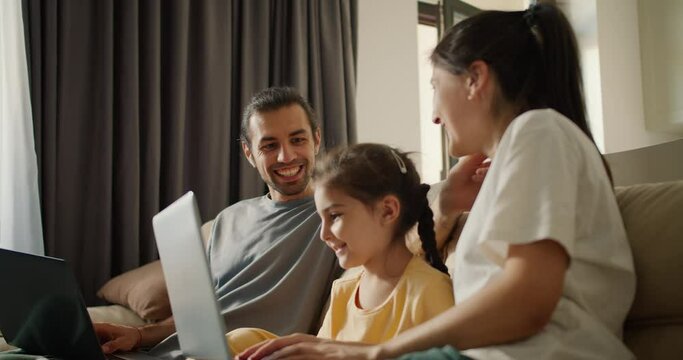 Family sitting on the sofa and looking at something on a gray laptop. A brunette girl in a white T-shirt, her daughter, a girl in a yellow dress, and a brunette man in a gray T-shirt are sitting