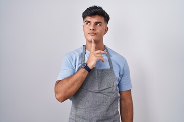 Hispanic young man wearing apron over white background thinking concentrated about doubt with...