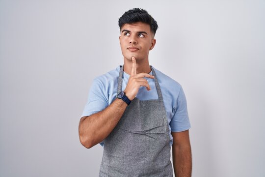 Hispanic young man wearing apron over white background thinking concentrated about doubt with finger on chin and looking up wondering