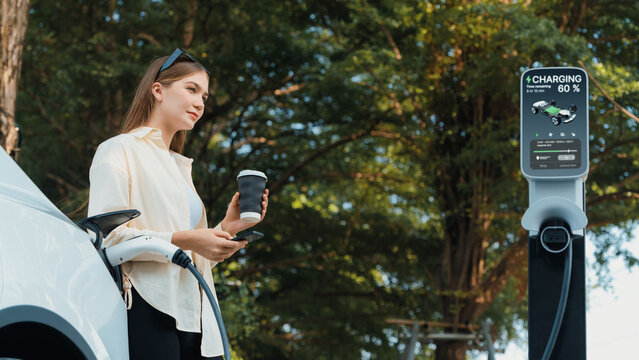 Young woman with coffee cup and sustainable urban commute with EV electric car recharging at outdoor cafe in springtime garden, green city sustainability and environmental friendly EV car. Expedient