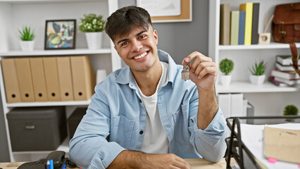 Smiling young hispanic real state agent in the office, celebrating success as he holds the key to a...