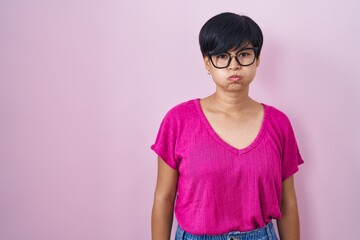 Young asian woman with short hair standing over pink background puffing cheeks with funny face. mouth inflated with air, crazy expression. - obrazy, fototapety, plakaty