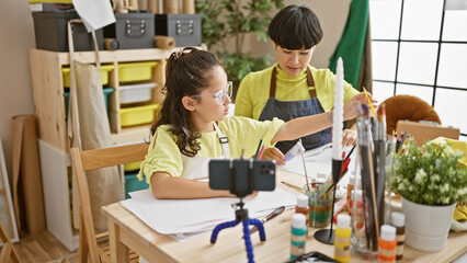 Cheerful student and her art teacher-artist engage in drawing lesson, recording video tutorial on notebook in cozy studio, fostering creativity together