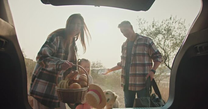 A middle-aged man with gray hair in a plaid shirt and his wife, a brunette girl in a green plaid shirt, together with their daughter, are packing things that are necessary for a picnic while driving