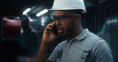 African American industrial worker talks by mobile phone, consults about settings of energy system....