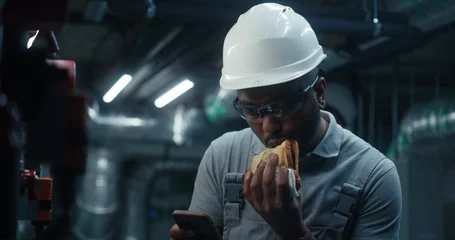  African American engineer eats sandwich and surfs the Internet on phone. Professional worker in uniform and protective hard hat having break working on industrial factory or energy facility. Close up. © Framestock