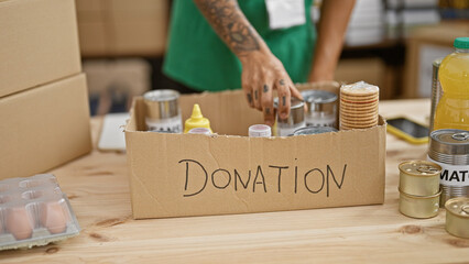 Tattooed, hispanic man's gracious hands volunteer, putting canned food donations in a cardboard box...