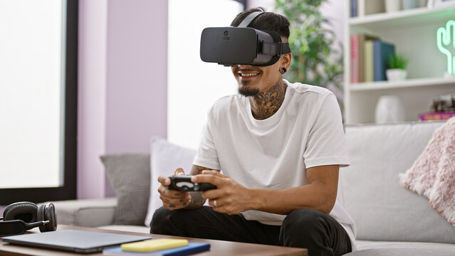 Confident young latin gamer happily using a joystick and vr glasses while playing intense video games in his comfortable living room