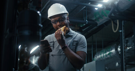 African American technician eats sandwich and surfs the Internet on mobile phone. Professional...