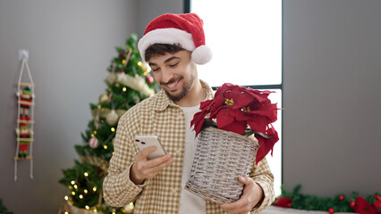 Young arab man using smartphone holding plant standing by christmas tree at home