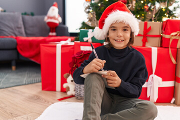 Obraz na płótnie Canvas Adorable hispanic boy writing santa claus letter sitting by christmas tree at home