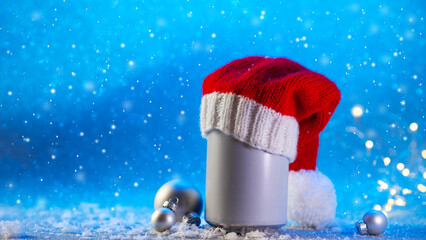A beer or soda can in a Santa Claus hat on a Christmas blue background with snow. Copy space