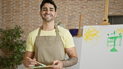 Young hispanic man artist smiling confident holding paintbrush and palette at art studio