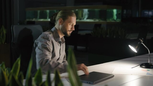 Overworked overtime working businessman manager man at night evening dark office finish end laptop work turn off light table lamp leaving workplace business male finishing job go away walking home