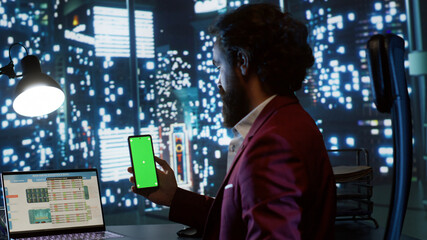 Qualified consultant operates on famous global company while looking at mobile device with greenscreen interface. In skyscraper office building, business leader checks isolated copyspace display.