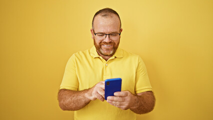 Cheerful bearded caucasian man with glasses, joyfully using smartphone, typing, sending happy texts over yellow isolated background