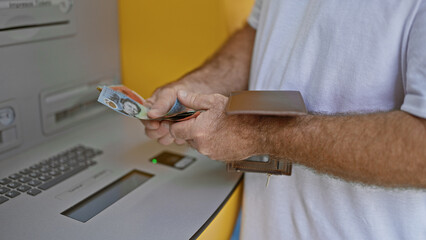 Obraz premium Handsome middle-aged caucasian man outside on urban street, captivating portrait of him holding wallet counting australian cash dollars at atm machine, engaging in banking transaction.