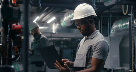 Professional African American inspector checks piping system on modern factory. Multiethnic team of technician specialists work on plant using laptop and tablet computers. Concept of heavy industry.